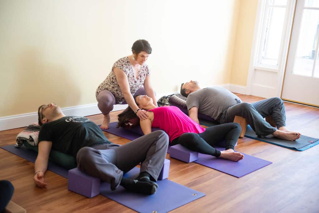 three people laying on purple mats in a yoga position with the instructor, michele, helping the person in the middle with positioning